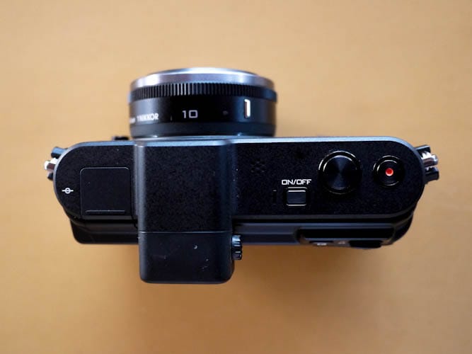 The top section of Nikon V1 is pretty clean, there are three buttons on the right - the shutter button, video record button and a button to switch the camera on and off, the viewfinder is placed at the center and the the hot shoe at the left.