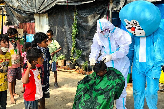 A barber wearing protective gear gives a haircut to a child next to a man dressed as comic character Doraemon in a Mumbai slum amid Covid-19 lockdown