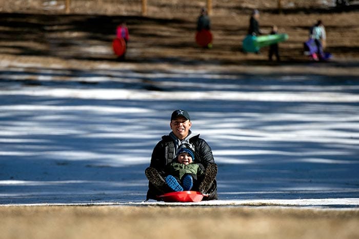 A man holding a young bo slide onto bare grass while sledding at Carter Park in Breckenridge, Colorado.